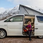 Guitarist sitting inside white toyota alphard campervan with open roof top box beside snowy mountain