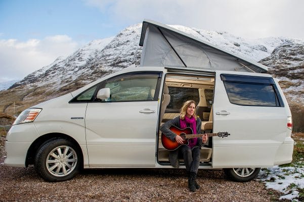 Guitarist sitting inside white toyota alphard campervan with open roof top box beside snowy mountain