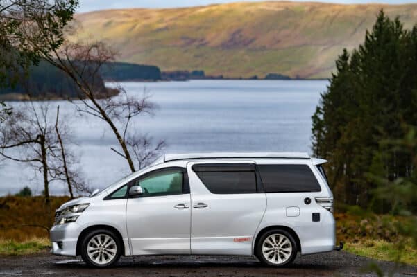 Nearside view of Toyota Alphard Connect II Campervan in mountain area with roof down next to loch and trees