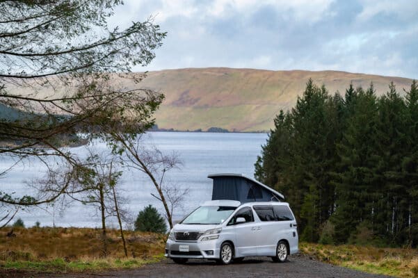 Nearside view of Toyota Alphard Connect II Campervan in mountain area with roof up next to loch and trees from distance