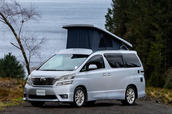 Nearside view of Toyota Alphard Connect II Campervan in mountain area with roof up next to loch and trees